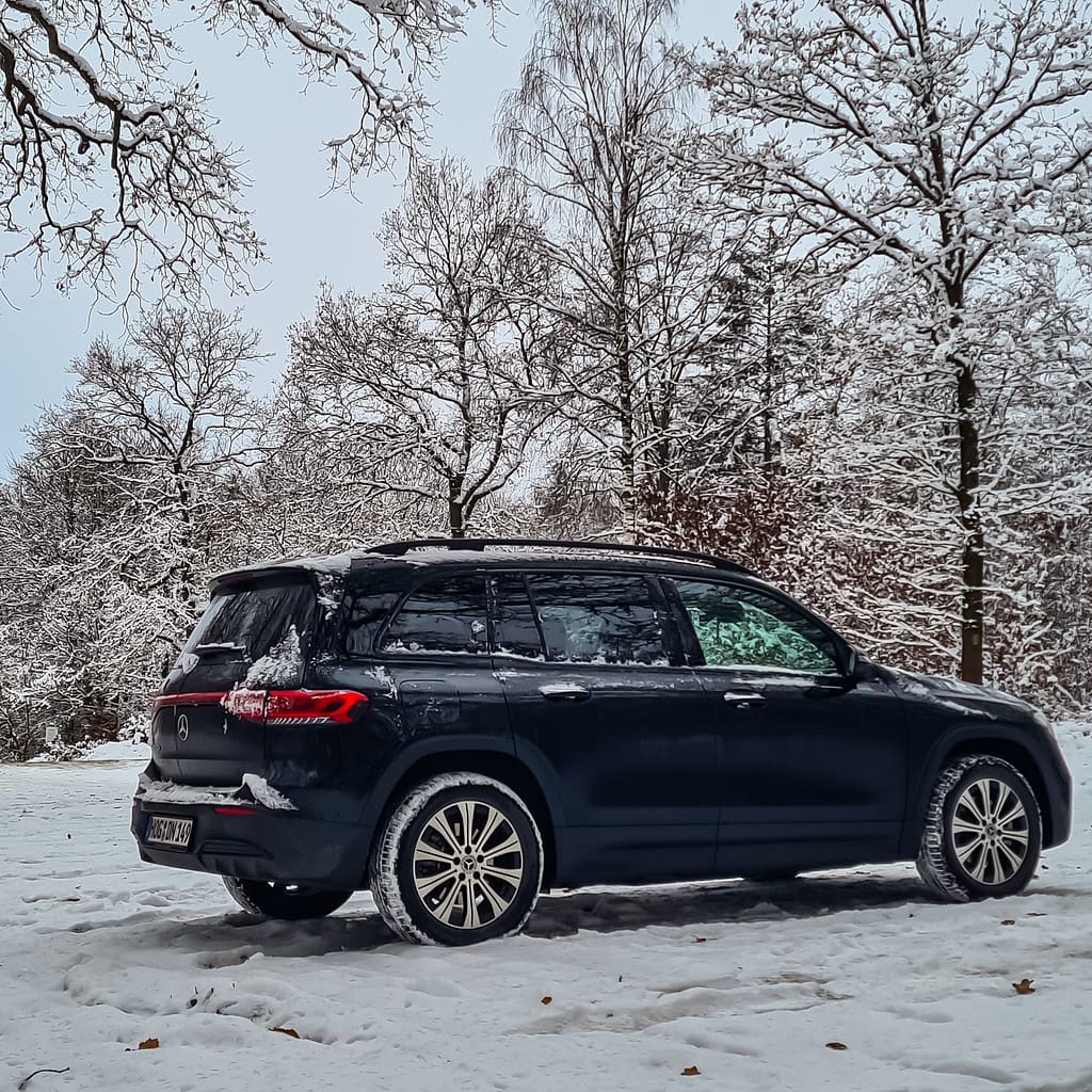 Dunkler Mercedes-Benz EQB X243 mit AMG Line in seitlicher Heckansicht im verschneiten Winterwald Bildtitel: Mercedes-Benz EQB (X243) mit AMG Line im Winter