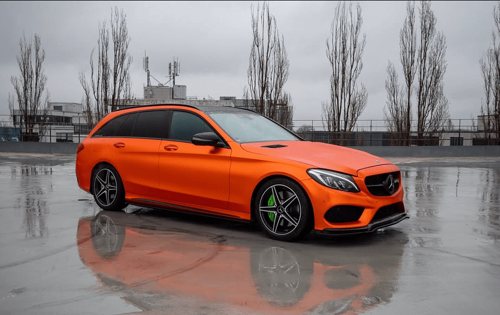 Orange foliertes Mercedes-Benz C 43 AMG T-Modell (S205) auf einem regennassen Parkdeck mit Spiegelungen auf dem Asphalt, schwarzem Dach und sportlichen AMG-Designelementen.