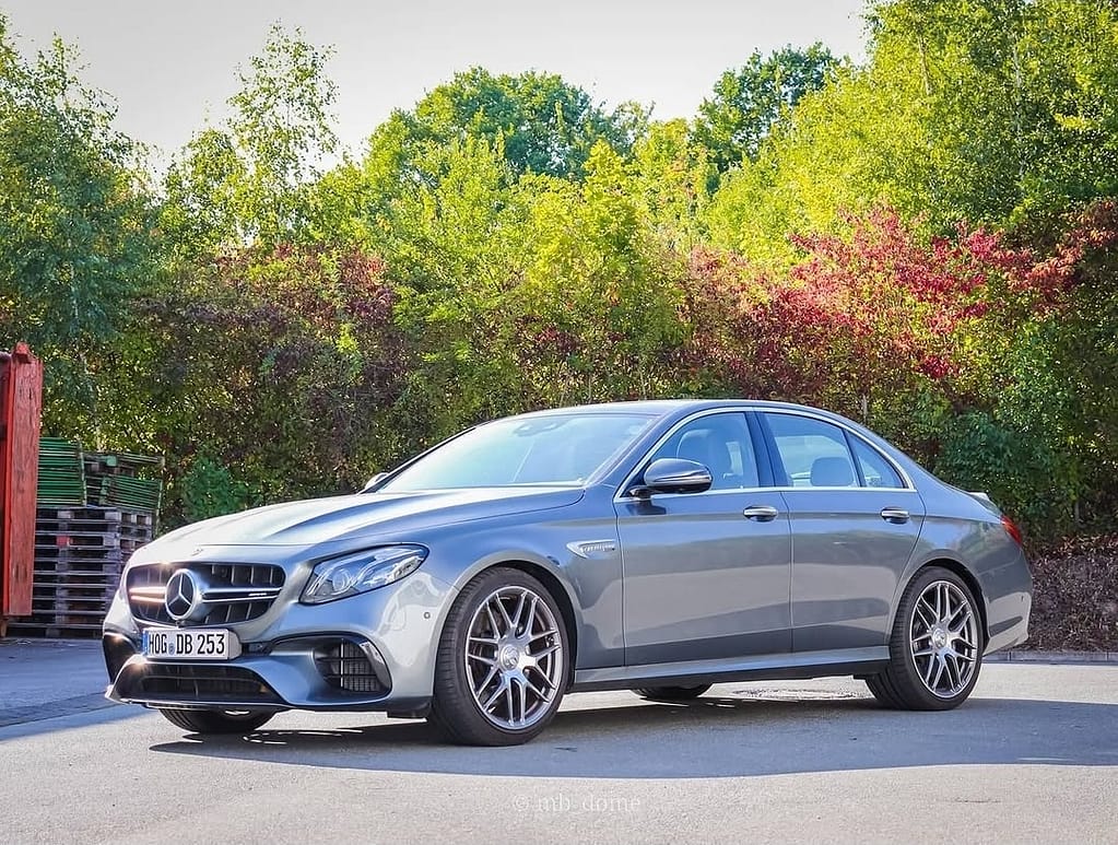 Mercedes-AMG E63 in seitlicher Frontperspektive auf einem Parkplatz mit grüner Vegetation und bunten Büschen im Hintergrund, sportliche AMG-Limousine mit markanten Felgen.