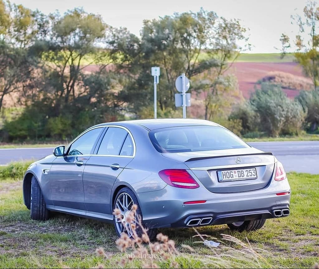 Mercedes-AMG E63 in Heck-Seitenansicht auf einer Wiese am Rand einer Landstraße mit Feldern und Bäumen im Hintergrund fotografiert