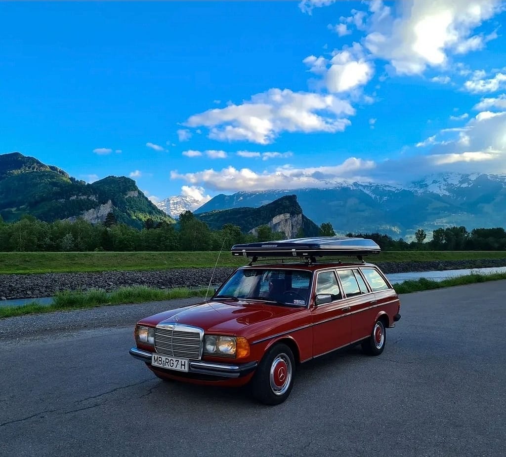 Roter Mercedes-Benz 240 TD S123 aus der Front-Seitenansicht vor einer eindrucksvollen Alpenkulisse