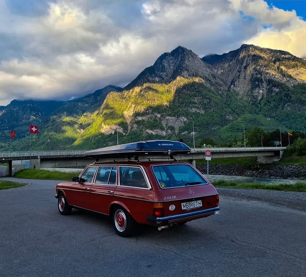 Roter Mercedes-Benz 240 TD der Baureihe S123 aus der Heck-Seitenansicht vor einer Bergkulisse