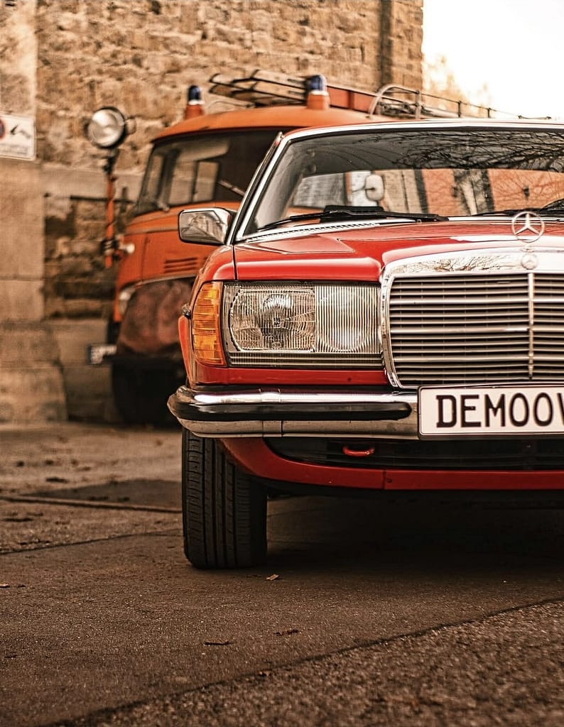 Rotes Mercedes-Benz C123 Coupé vor historischem Feuerwehrfahrzeug von Mercedes-Benz. Klassische Mercedes-Fahrzeuge vor historischer Architektur fotografiert bei warmem Abendlicht.