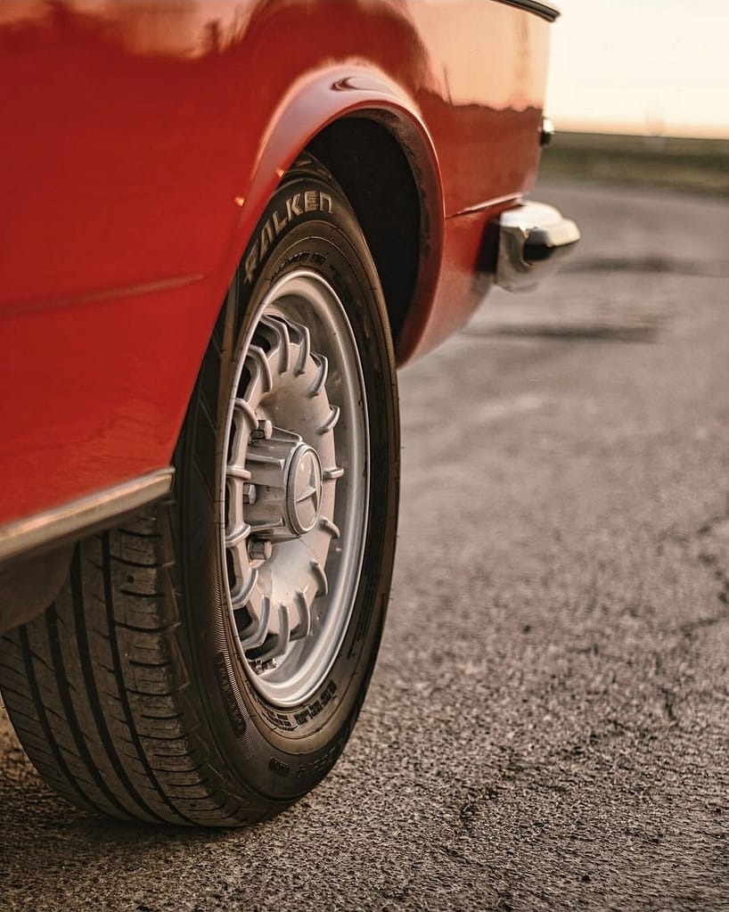 Detailaufnahme der originalen Mercedes-Benz Felge und des Reifens eines roten C123 Coupé bei Sonnenuntergang.