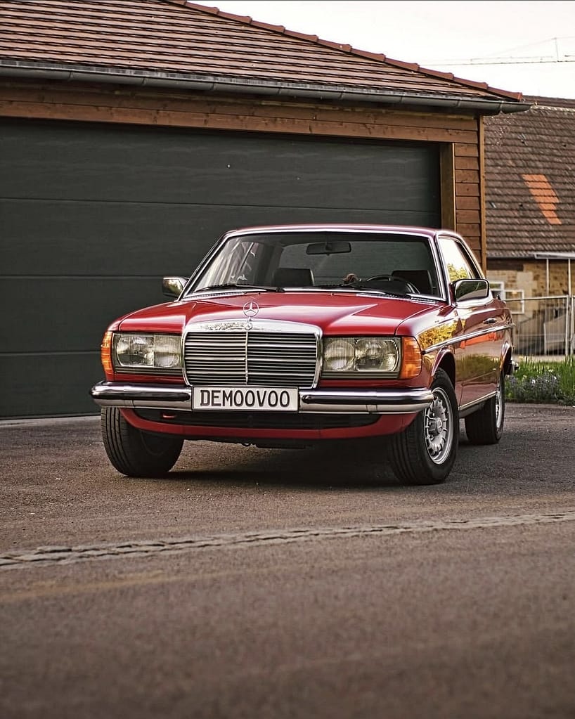 Mercedes-Benz C123 Coupé in roter Lackierung bei Abendlicht vor einer Garage fotografiert. Klassischer Mercedes-Benz Oldtimer mit Chromgrill und zeitlosem Coupé-Design.