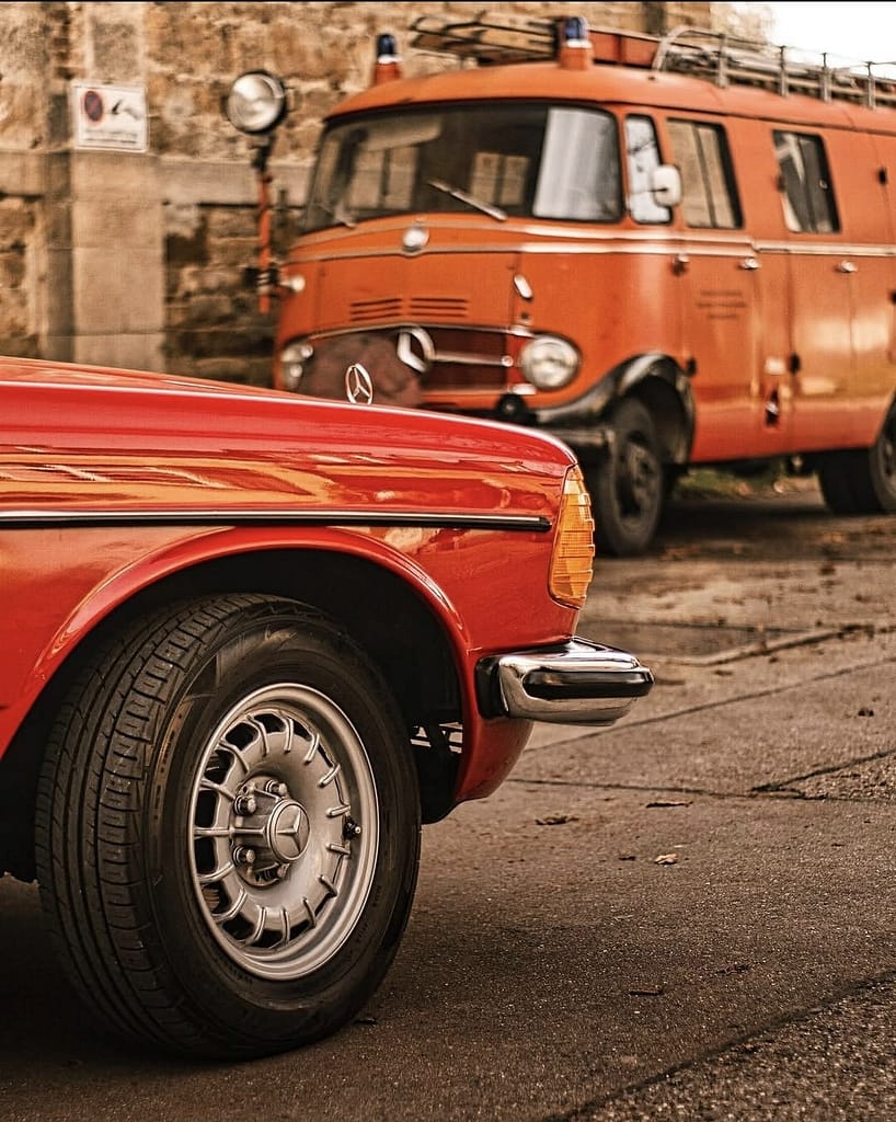Rotes Mercedes-Benz C123 Coupé vor historischem Feuerwehrfahrzeug von Mercedes-Benz. Klassische Mercedes-Fahrzeuge vor historischer Architektur fotografiert bei warmem Abendlicht.