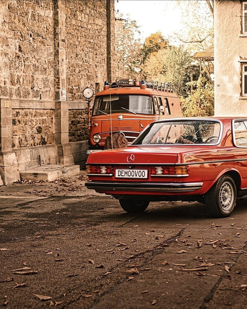 Rotes Mercedes-Benz C123 Coupé vor historischem Feuerwehrfahrzeug von Mercedes-Benz. Klassische Mercedes-Fahrzeuge vor historischer Architektur fotografiert bei warmem Abendlicht.