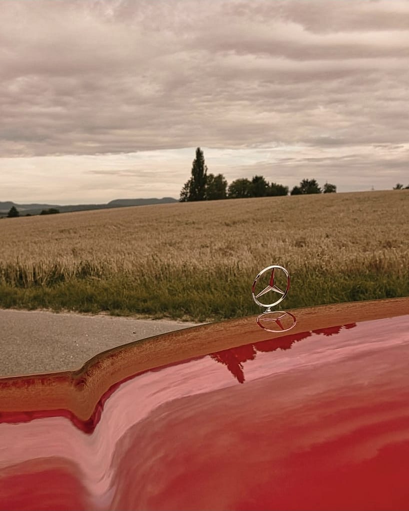Mercedes-Benz C123 Coupé in klassischem Rot auf einer ländlichen Straße vor Getreidefeldern und dramatischem Wolkenhimmel – hochwertige Automotive Photography von MB DOME.