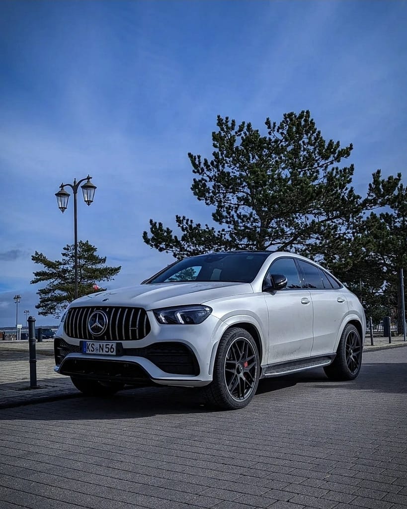 Mercedes-AMG GLE 53 Coupé (C167) in Polarweiß mit Panamericana-Grill am Hafen fotografiert. Front- und Seitenansicht des sportlichen AMG SUV-Coupés vor maritimer Kulisse unter blauem Himmel.