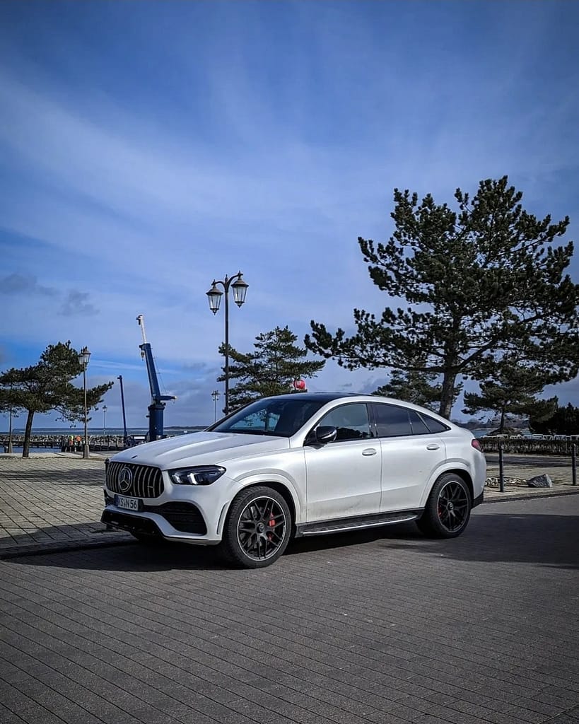 Mercedes-AMG GLE 53 Coupé (C167) in Polarweiß mit Panamericana-Grill am Hafen fotografiert. Front- und Seitenansicht des sportlichen AMG SUV-Coupés vor maritimer Kulisse unter blauem Himmel.