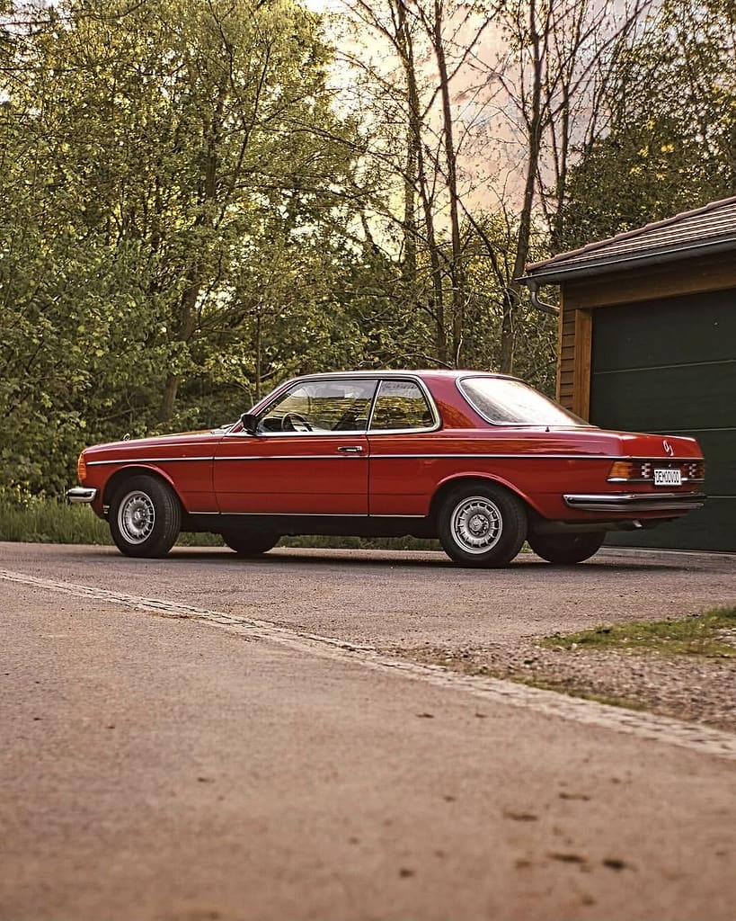 Mercedes-Benz C123 Coupé in roter Lackierung bei Abendlicht vor einer Garage fotografiert. Klassischer Mercedes-Benz Oldtimer mit Chromgrill und zeitlosem Coupé-Design.