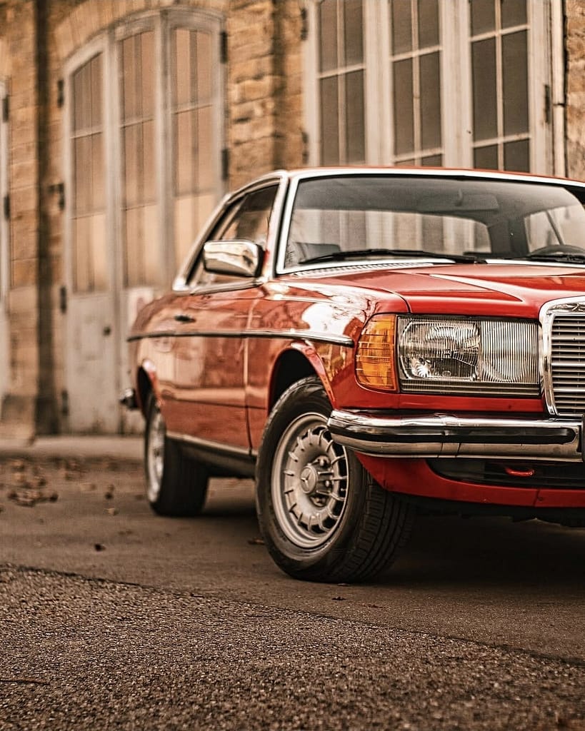 Rotes Mercedes-Benz C123 Coupé vor historischem Feuerwehrfahrzeug von Mercedes-Benz. Klassische Mercedes-Fahrzeuge vor historischer Architektur fotografiert bei warmem Abendlicht.