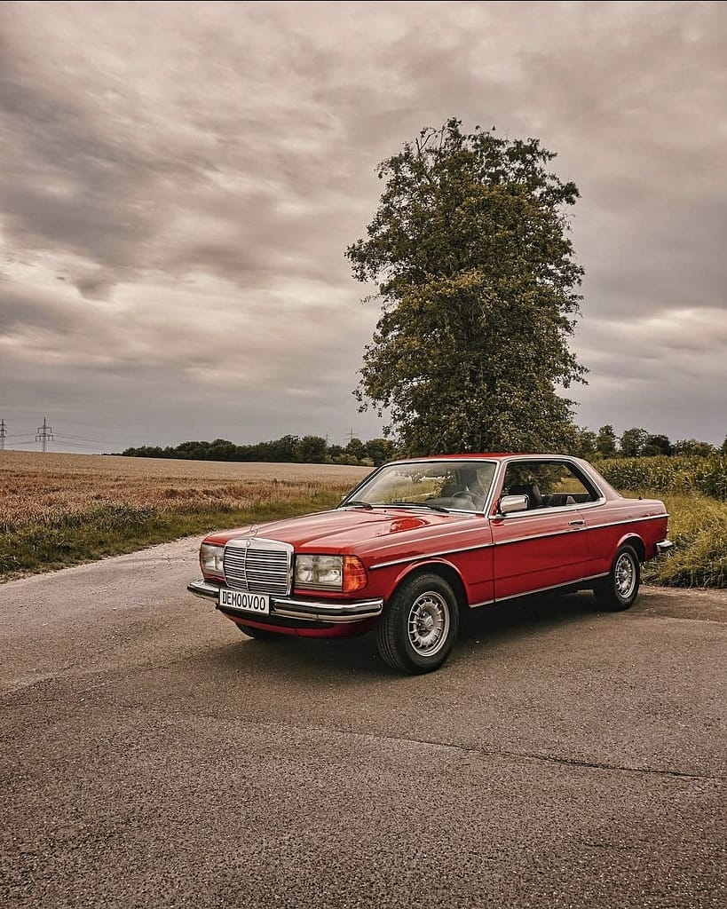 Mercedes-Benz C123 Coupé in klassischem Rot auf einer ländlichen Straße vor Getreidefeldern und dramatischem Wolkenhimmel – hochwertige Automotive Photography von MB DOME.