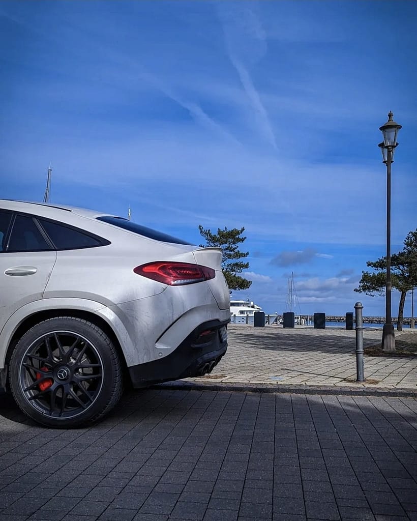 Mercedes-AMG GLE 53 Coupé (C167) in Polarweiß mit Panamericana-Grill am Hafen fotografiert. Front- und Seitenansicht des sportlichen AMG SUV-Coupés vor maritimer Kulisse unter blauem Himmel.