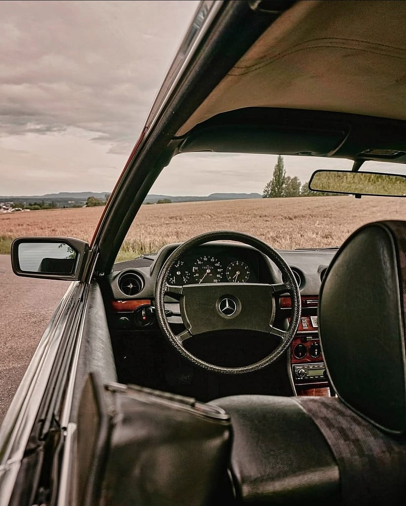 Mercedes-Benz C123 Coupé in klassischem Rot auf einer ländlichen Straße vor Getreidefeldern und dramatischem Wolkenhimmel – hochwertige Automotive Photography von MB DOME.