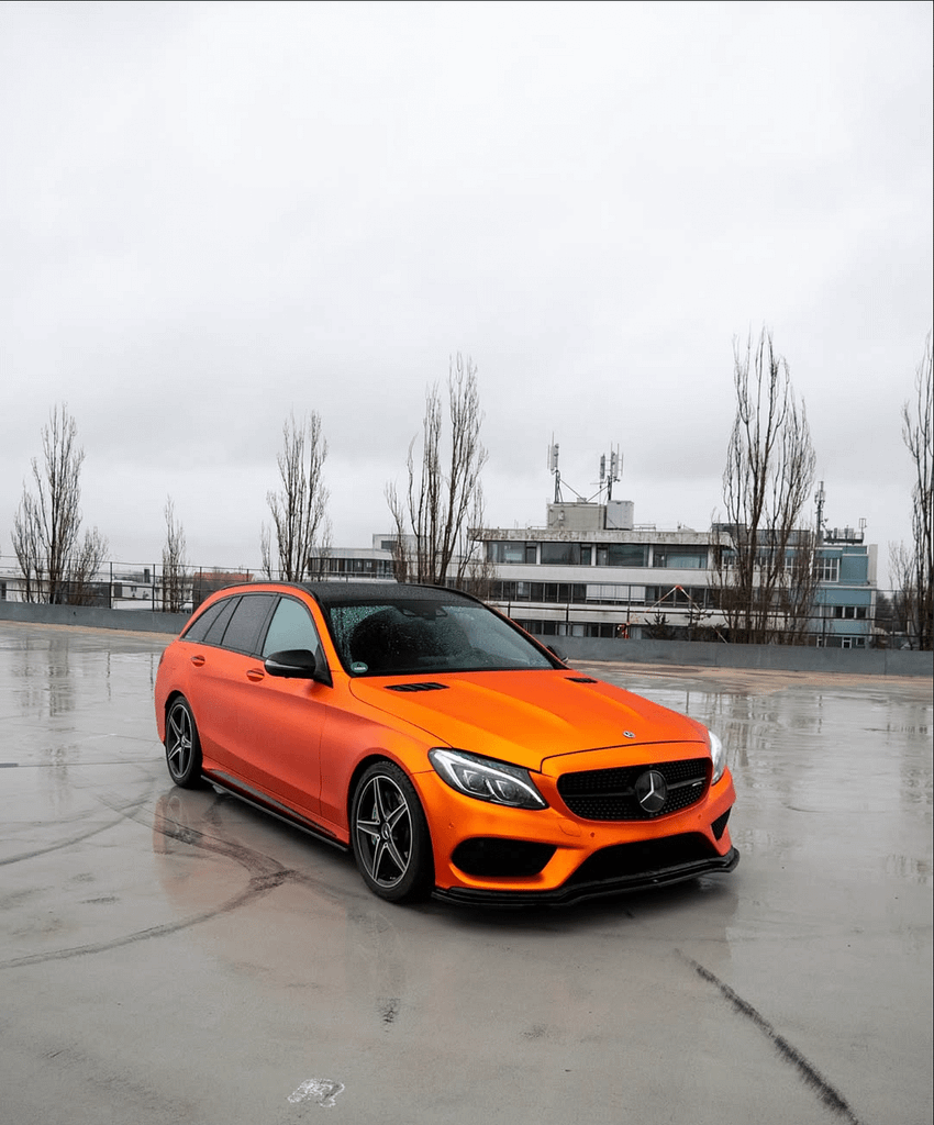 Orange foliertes Mercedes-Benz C 43 AMG T-Modell (S205) auf einem regennassen Parkdeck mit Spiegelungen auf dem Asphalt, schwarzem Dach und sportlichen AMG-Designelementen.