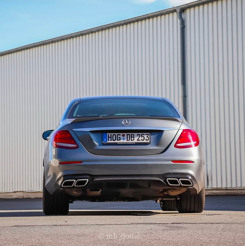 Heckansicht eines grauen Mercedes-AMG E63 mit vier AMG-Endrohren und LED-Rückleuchten vor einer hellen Industriehalle auf einem Parkplatz.