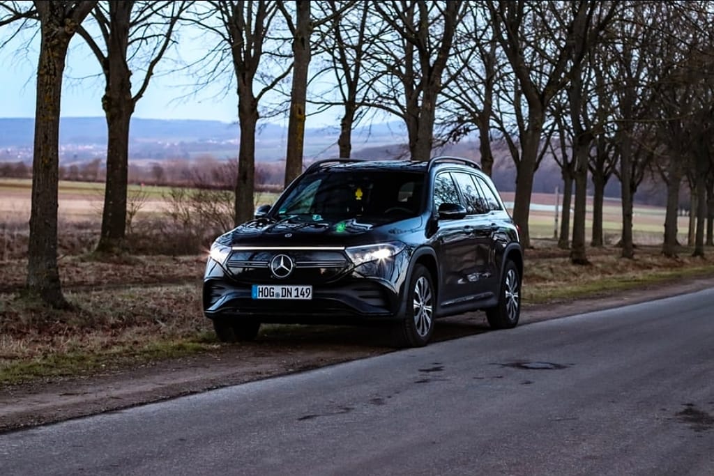 Mercedes-Benz EQB in Schwarz am Straßenrand zwischen Bäumen bei Abendlicht mit eingeschalteten Frontscheinwerfern fotografiert