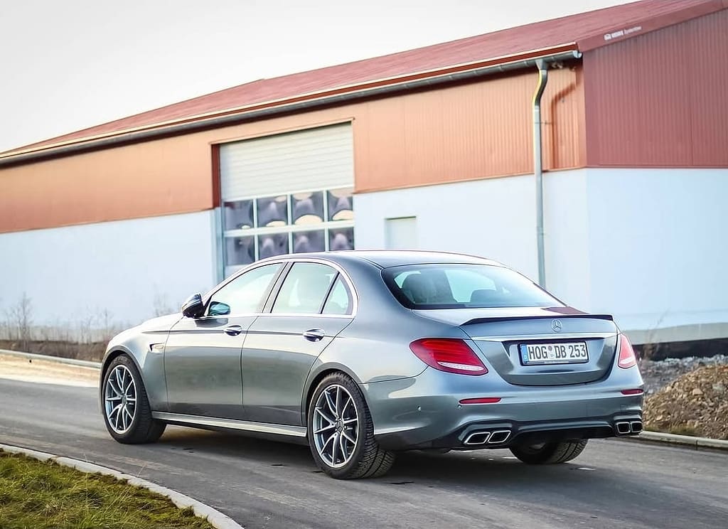Mercedes-AMG E63 in Heck-Seitenansicht auf einer Straße vor moderner Industriehalle mit sichtbarer AMG-Heckpartie und sportlichen Doppelendrohren.