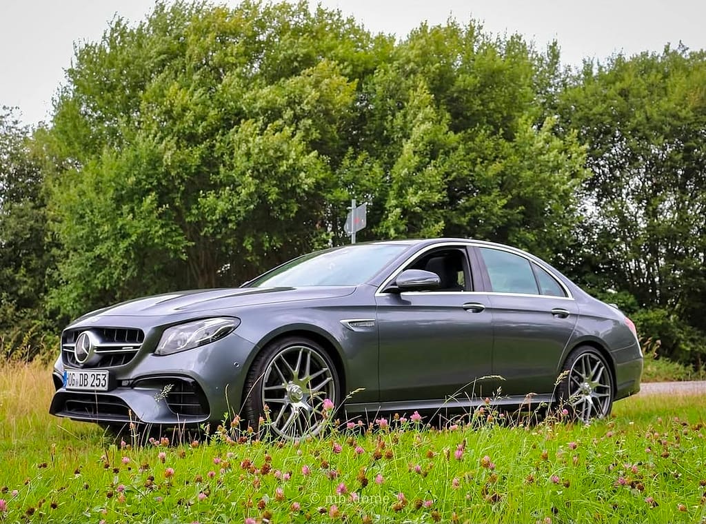 Mercedes-AMG E63 in Seitenansicht auf einer Wiese mit Blumen vor grüner Baumkulisse, sportliche AMG-Limousine mit markanten Felgen.