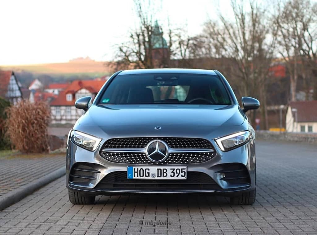 Frontansicht einer grauen Mercedes-Benz A-Klasse mit Diamantgrill und Mercedes-Stern auf einer gepflasterten Straße vor einer ruhigen Ortschaft im Hintergrund.