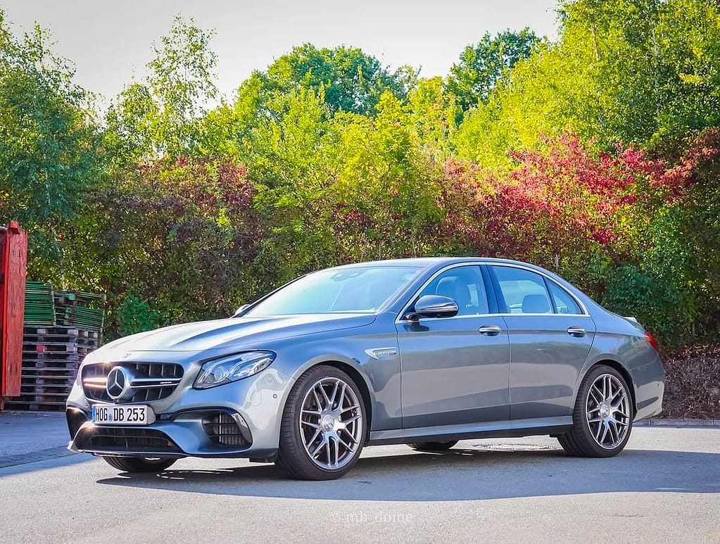 Mercedes-AMG E63 in seitlicher Frontperspektive auf einem Parkplatz mit grüner Vegetation und bunten Büschen im Hintergrund, sportliche AMG-Limousine mit markanten Felgen.