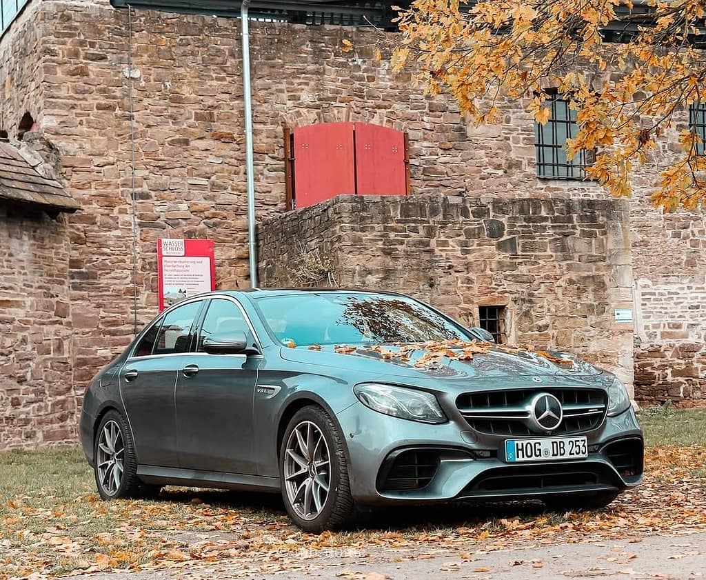 Mercedes-AMG E63 in Front-Seitenansicht mit Herbstlaub auf der Motorhaube vor historischer Natursteinmauer mit rotem Tor