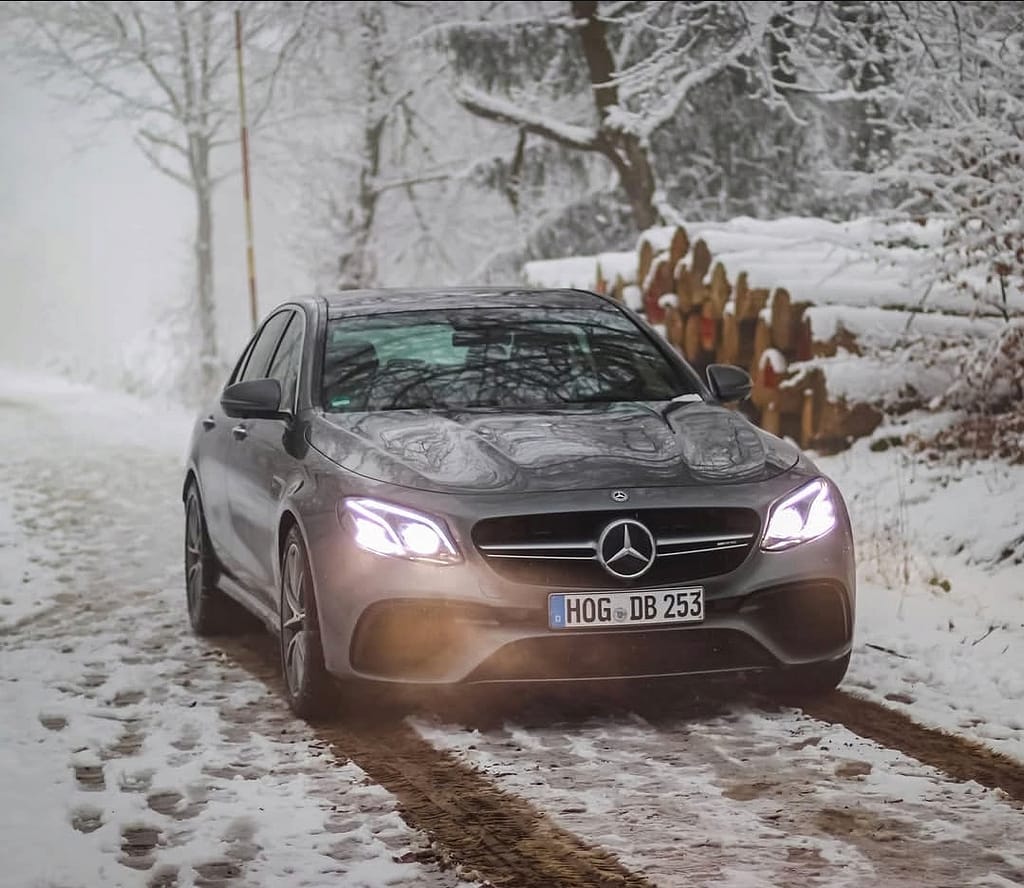 Mercedes-AMG E63 in Front-Seitenansicht auf verschneitem Waldweg mit eingeschalteten LED-Scheinwerfern fotografiert