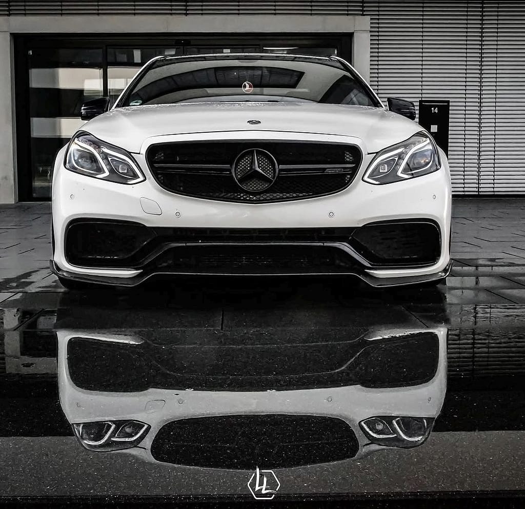 Frontansicht eines weißen Mercedes-Benz E63 AMG W212 mit Spiegelung auf nasser Straße vor moderner Architektur