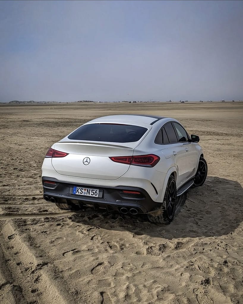 Weißes Mercedes-AMG GLE 53 Coupé C167 am Strand mit sportlicher Heckansicht und schwarzen AMG Felgen.
