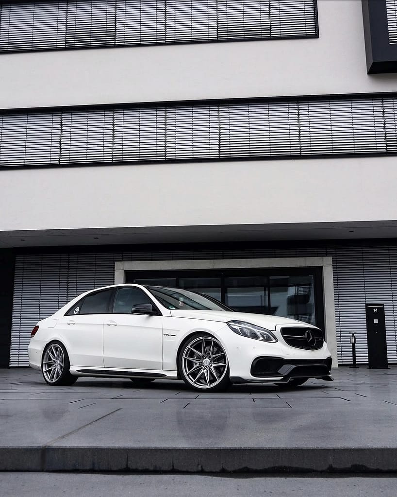Weißer Mercedes-Benz E63 AMG W212 mit Tieferlegung und großen Felgen vor moderner schwarzer und weißer Architektur