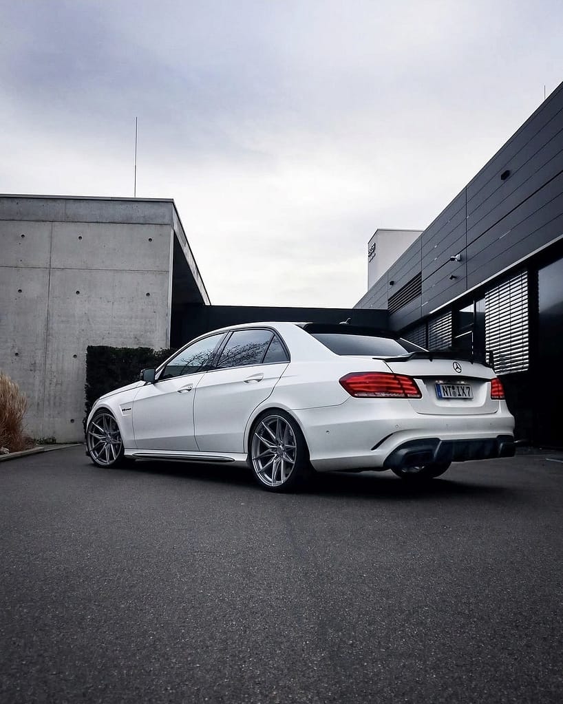 Weißer Mercedes-Benz E63 AMG W212 mit großen Felgen und Carbon-Heckspoiler vor moderner Architektur bei nasser Straße