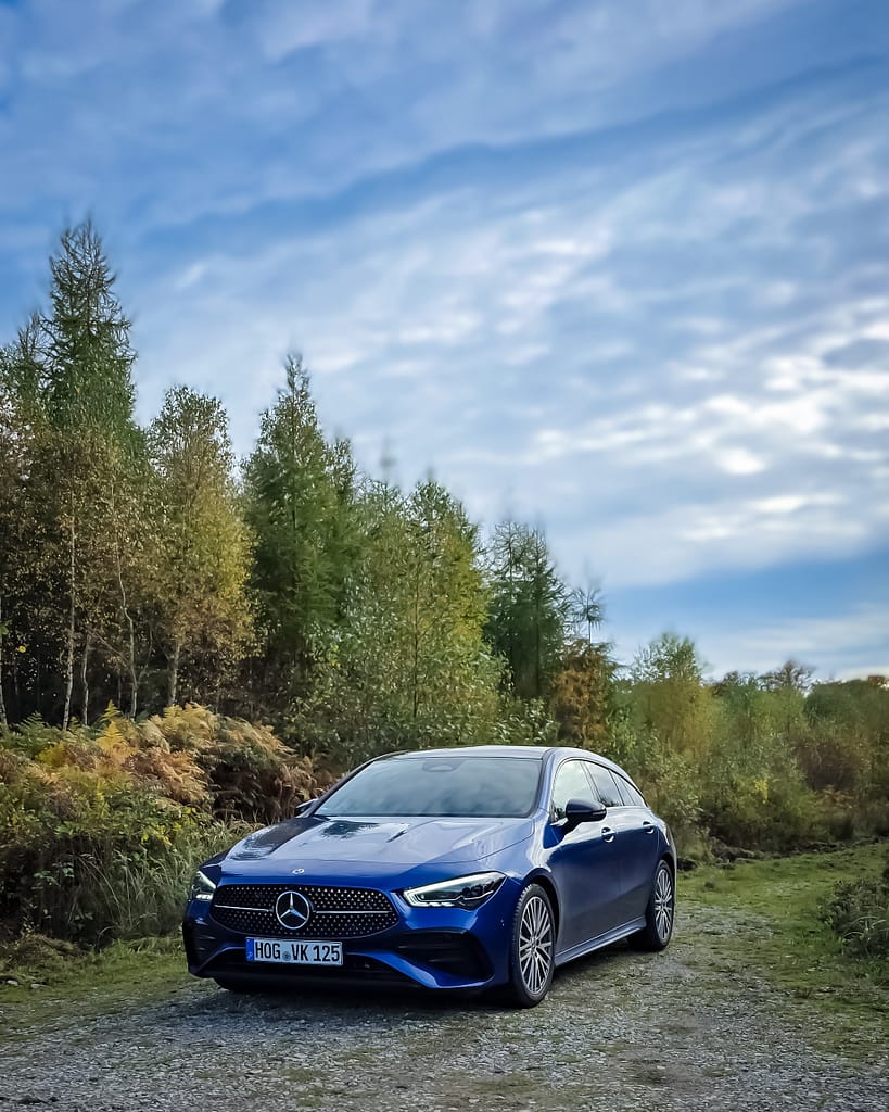 Blauer Mercedes-Benz CLA Shooting Brake auf einem Waldweg in Front-Seitenperspektive, umgeben von grüner Natur und Bäumen unter einem bewölkten Himmel.
