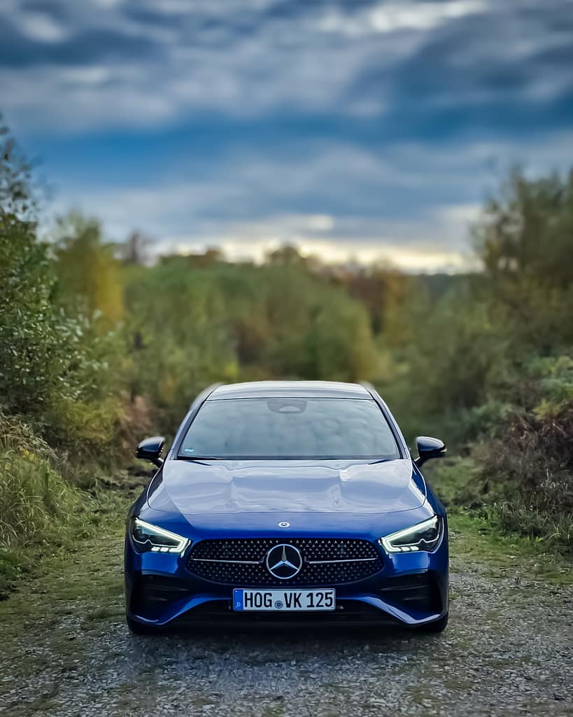 Frontansicht eines blauen Mercedes-Benz CLA auf einem schmalen Waldweg mit grünem Naturhintergrund und dramatischem Himmel, moderne Limousine mit LED-Scheinwerfern und Diamantgrill.