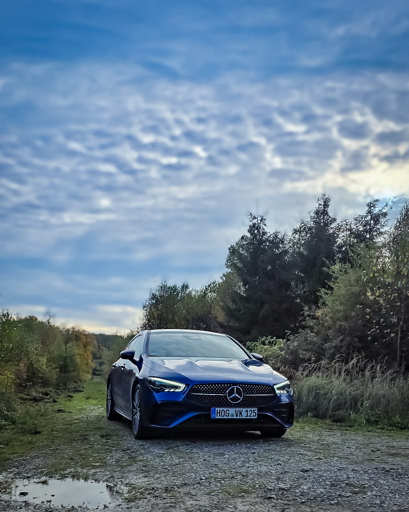 Blauer Mercedes-Benz CLA auf einem schmalen Waldweg unter einem dramatischen Wolkenhimmel, Frontansicht der modernen Mercedes-Limousine mit Diamantgrill und LED-Scheinwerfern.
