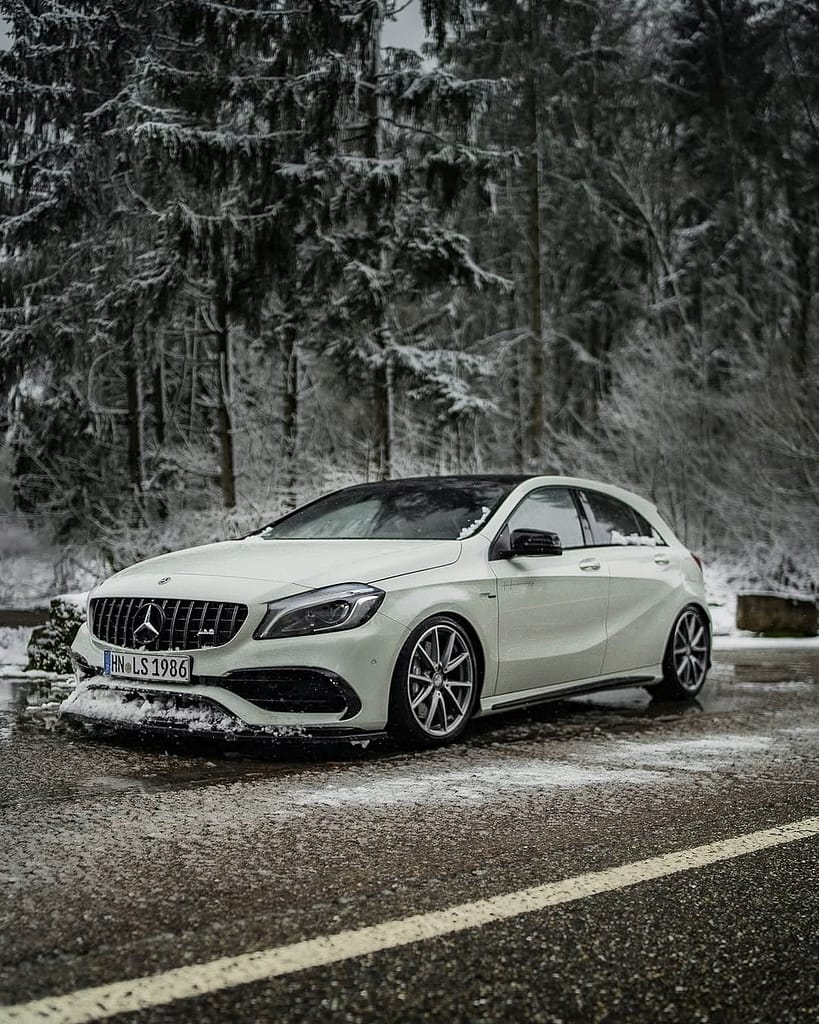Weißer Mercedes-AMG A45 W176 auf verschneiter Straße im Winterwald mit sportlicher Frontansicht und AMG Panamericana Grill
